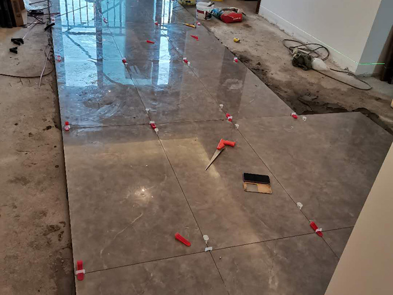 客廳地磚鋪貼：地磚鋪貼前，應將地面清掃干凈，尤其是凝結后的混凝土，否則，地磚鋪貼后會出現翹邊、空鼓等現象，然后再灑水潤濕地面。地面充分濕潤，但不能有積水，然后將3:1混合好的沙子水泥鋪在地面上，進行基層處理。在地磚背面均勻抹水泥砂漿作為粘合層，厚度不低于1cm，然后鋪在剛才試鋪的位置上，用橡皮錘輕敲地磚飾面直至密實平整達到要求。地磚鋪完后24h進行清理勾縫，勾縫前應先將地磚縫隙內雜質擦凈，用專用填縫劑勾縫，完工后（一般宜在24h之后）再用棉紗等物對地磚表面進行清理。