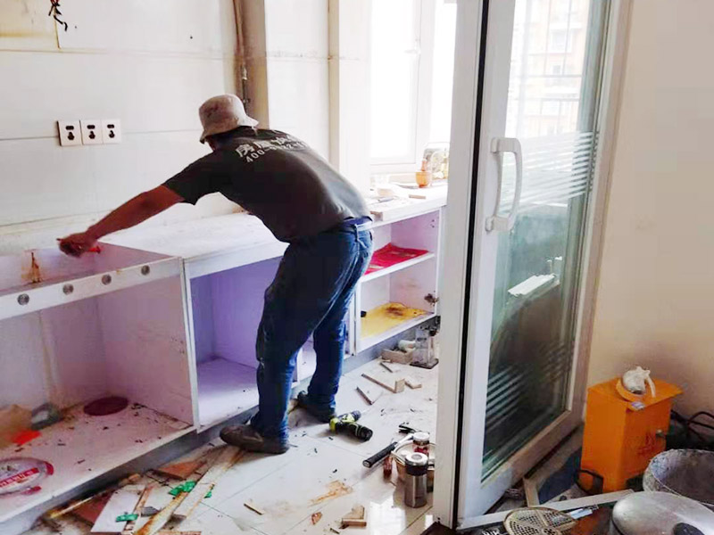 廚房櫥柜安裝：清理：對廚房作防護措施，掛擋布、廚房墻面等做簡單清理，以便準確測量地面水平。櫥柜安裝一般先從地柜開始。把櫥柜外包裝拆開后。在櫥柜的邊緣貼上防火膜封邊。在下柜底部安裝上支腳，根據地面水平高度調整櫥柜。 櫥柜安裝需要找出基準點，L型廚房從直角處向兩邊延伸，U型廚房則先將中間的一字型柜體放整齊后，再從兩個直角處向兩邊站看，這樣可以避免出現縫隙。櫥柜框架安裝牢固定位后，需要在櫥柜背面安裝背板。一般安裝下水都會現場開孔，工人會用專業的打孔器按照管道大小進行打孔，打孔的直徑應比管道大3-4毫米，并且打孔后要將開孔部分用密封條封邊，防止邊緣滲水木材變形。